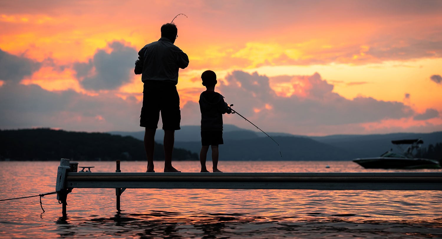 A father and child fishing together on a dock at sunset, capturing a peaceful outdoor moment with water and sky in the background.