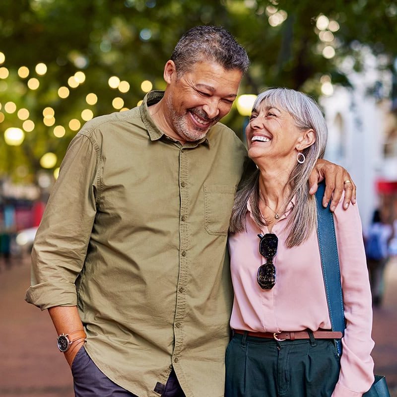 A cheerful elderly couple walking together in a lively outdoor setting with string lights, smiling and sharing a joyful moment, representing community and well-being.